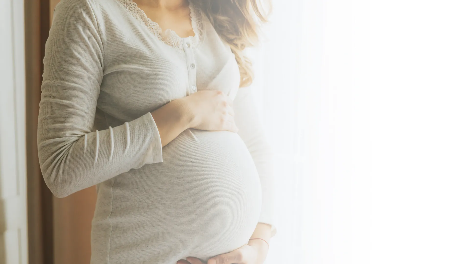 Gestante segurando a barriga próximo à janela, em luz suave